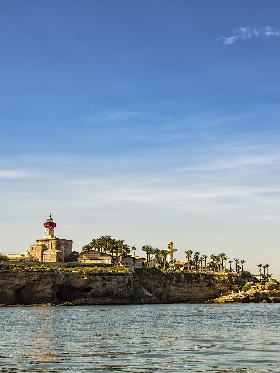 an image of a sicilian coast with a lighthouse and a calm sea underneath. on the coast there are various palm trees.